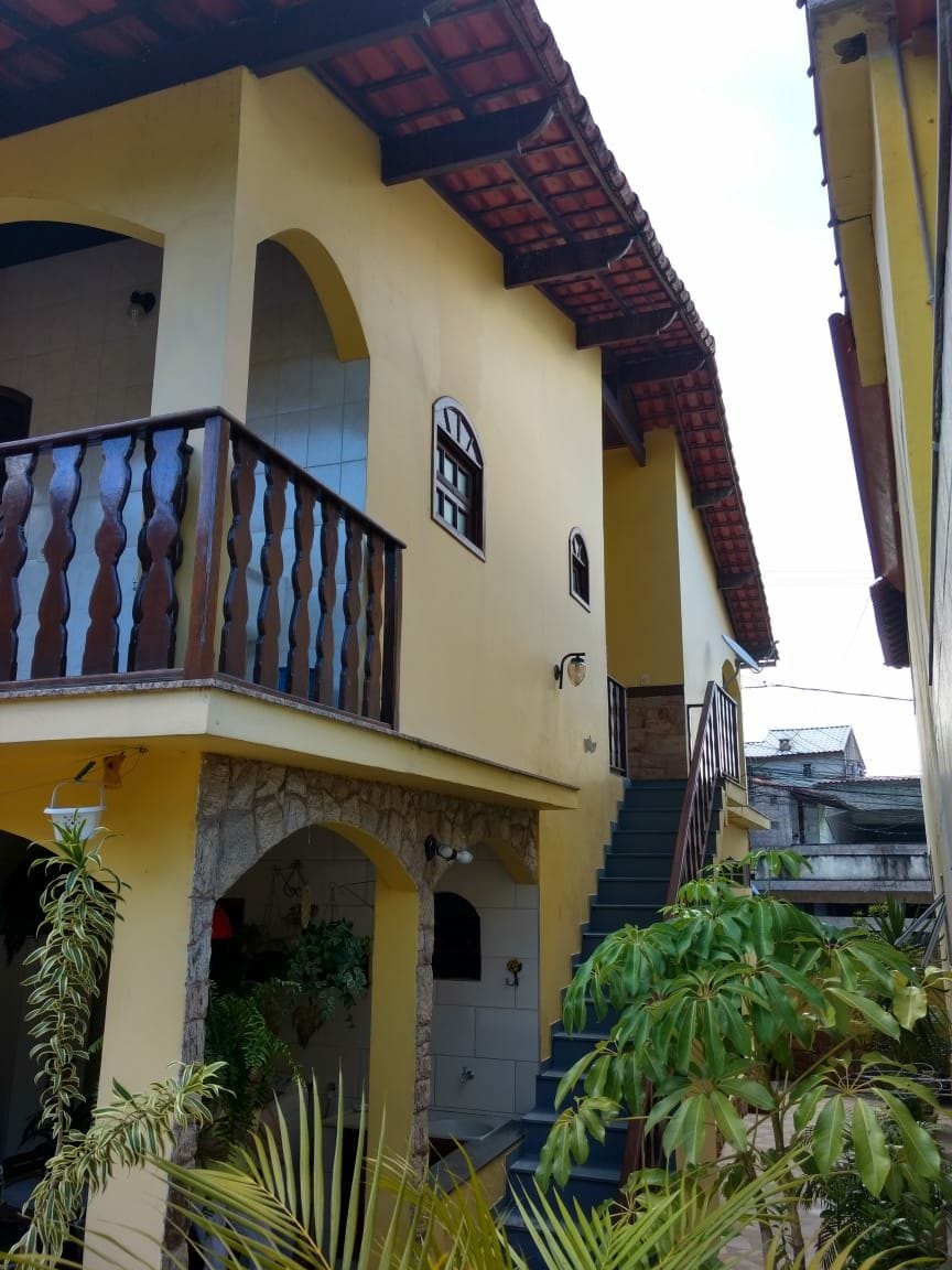 The exterior of a two-story house is shown, featuring a bright yellow facade and wooden balcony railings. Stairs lead to the entrance, framed by arches. Lush greenery includes potted plants near the base, enhancing the inviting appearance of the entryway.