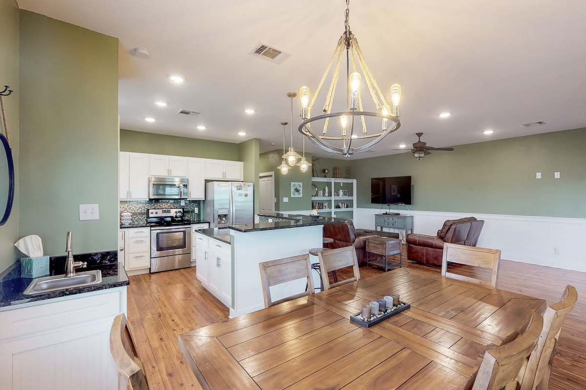 An open-concept living area features a dining table with wooden chairs, paired with a modern kitchen equipped with stainless steel appliances. Soft lighting from a large chandelier illuminates the space, which includes a comfortable seating area with plush sofas and a television.