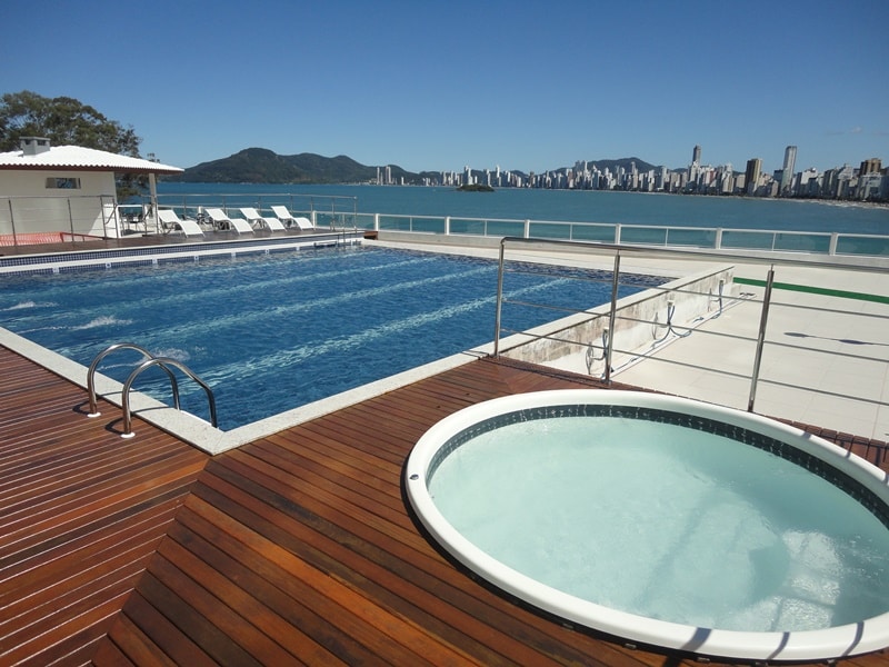 A large swimming pool is surrounded by a wooden deck and sun loungers, offering a clear view of the skyline. A separate circular hot tub sits adjacent to the pool, inviting relaxation, while the calm water reflects the sunny sky.