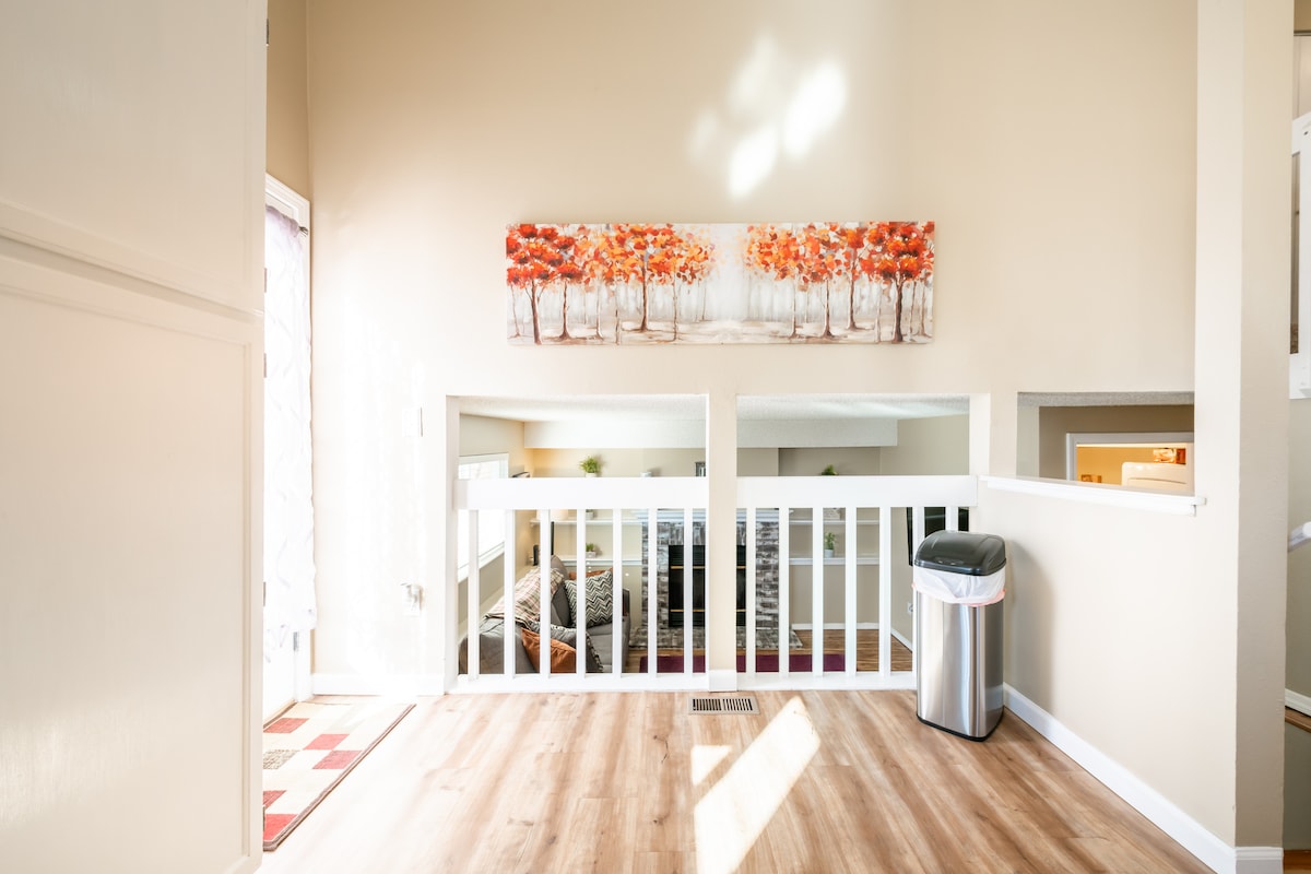 A bright and airy entryway features light-colored walls complemented by warm wood flooring. A painting of autumn trees hangs above a railing, which overlooks a cozy living area. A stylish trash bin is positioned nearby, contributing to the home's modern design.