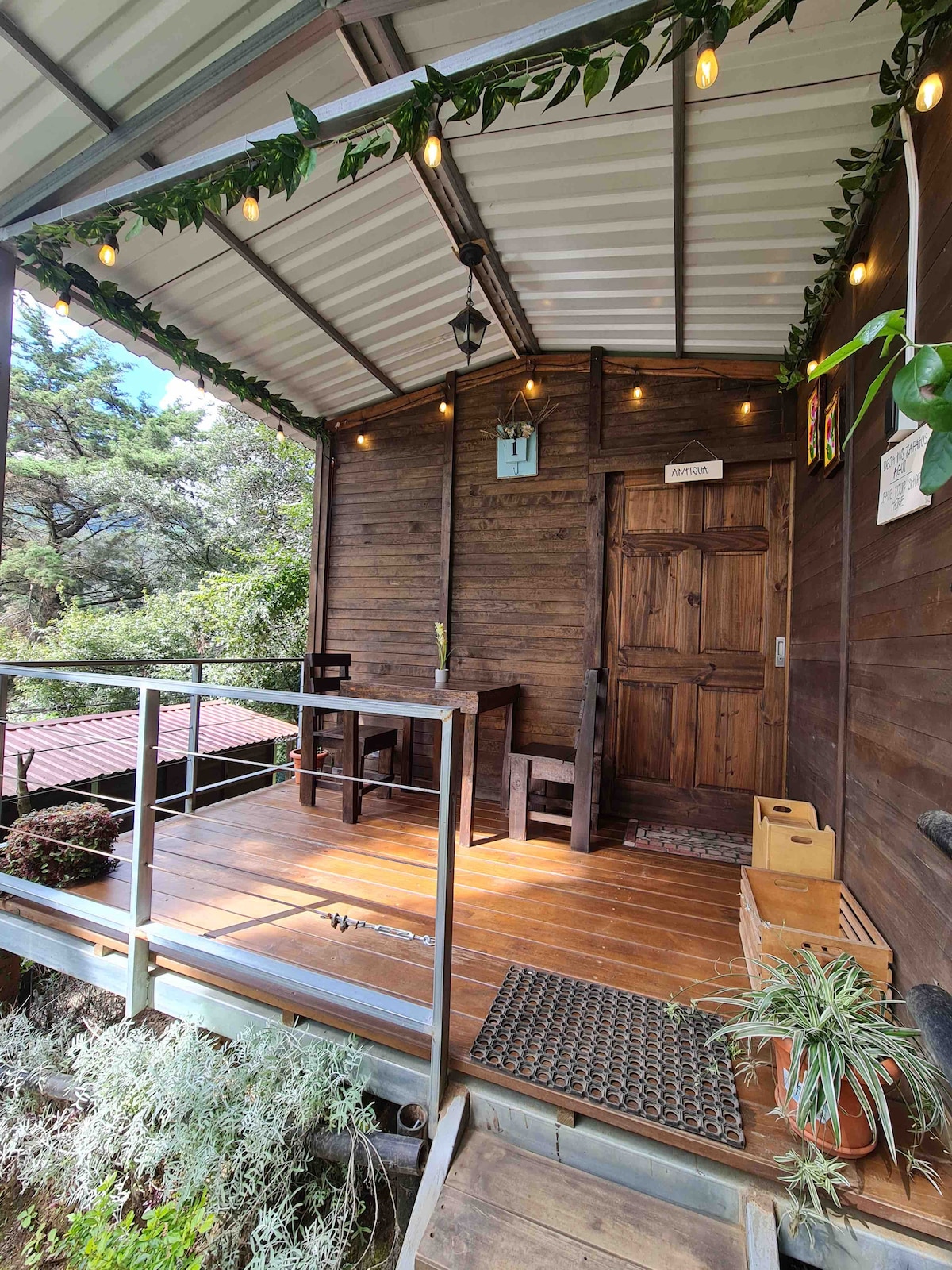 A welcoming entrance features a wooden door and decorative string lights above. Two chairs and a small table are situated on the covered porch, surrounded by greenery. A small mat lies at the entrance, contributing to the rustic charm of the cabin.