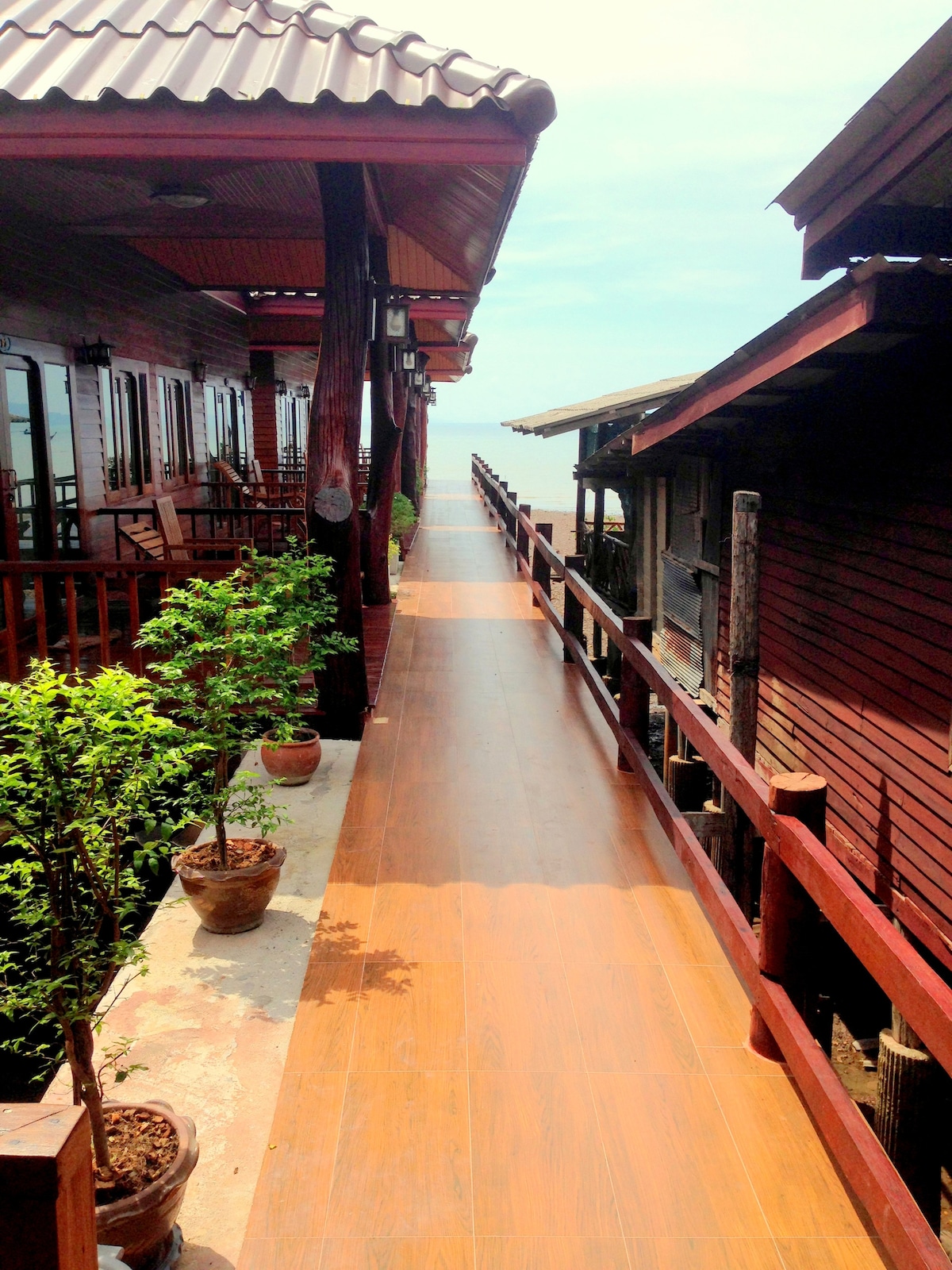 A wooden walkway extends between traditional wooden houses, bordered by potted plants. The sturdy railings provide a sense of safety and structure. Sunlight casts a warm glow on the smooth surface, and a clear sky is visible in the background.