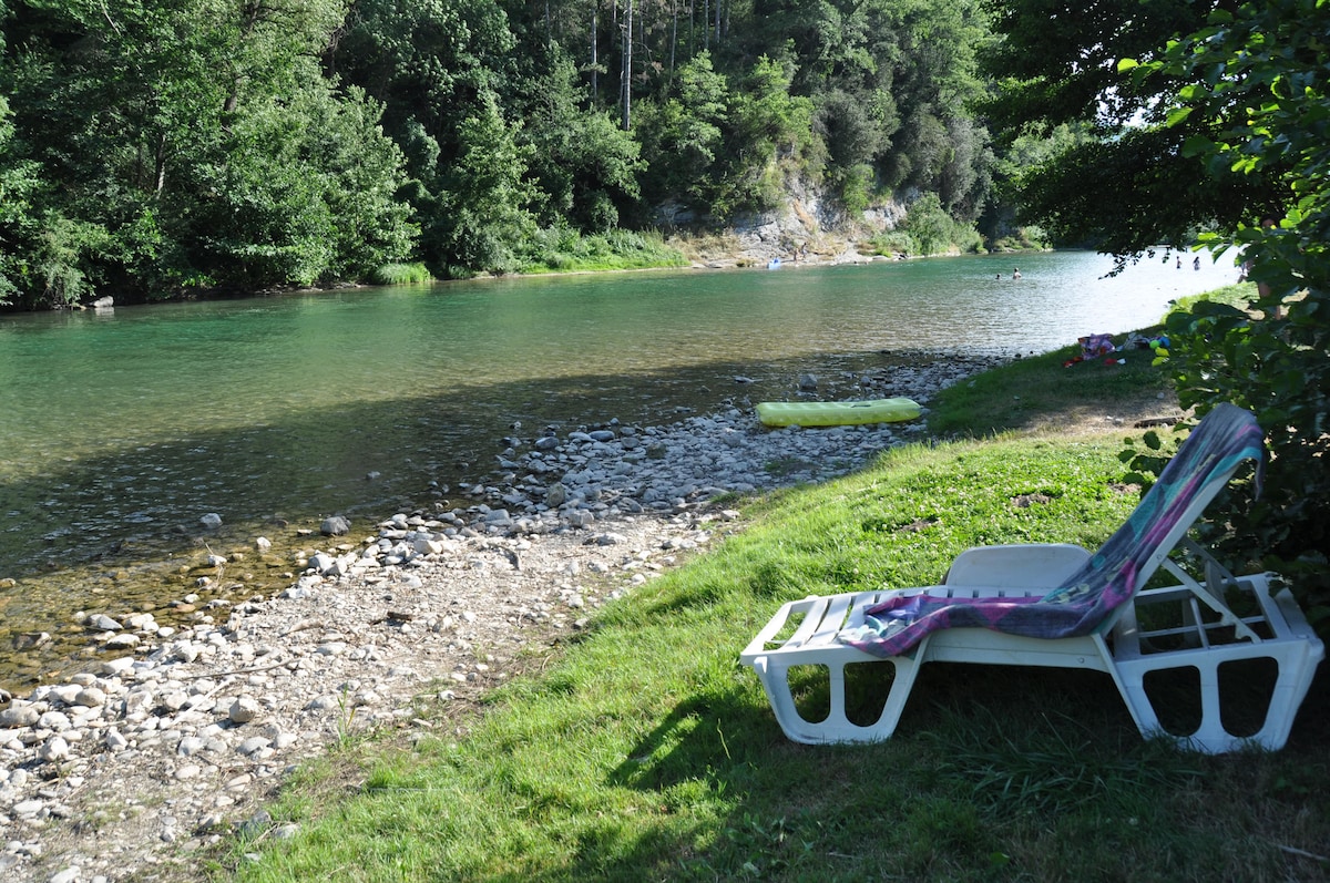 A sunlit area along a tranquil riverbank features a lounge chair placed on the grass, overlooking clear, calm waters. Smooth stones line the water's edge, with lush greenery providing shade. A yellow float is visible in the water, suggesting a relaxing outdoor environment.