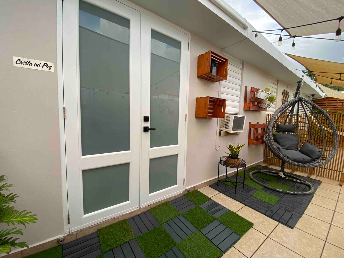 The entrance to Casita mi Paz features double glass doors framed by a beige wall. A hanging chair is positioned on a patterned tiled floor, complemented by decorative planters. Small wall-mounted shelves hold various items, while a window air conditioner is nearby.