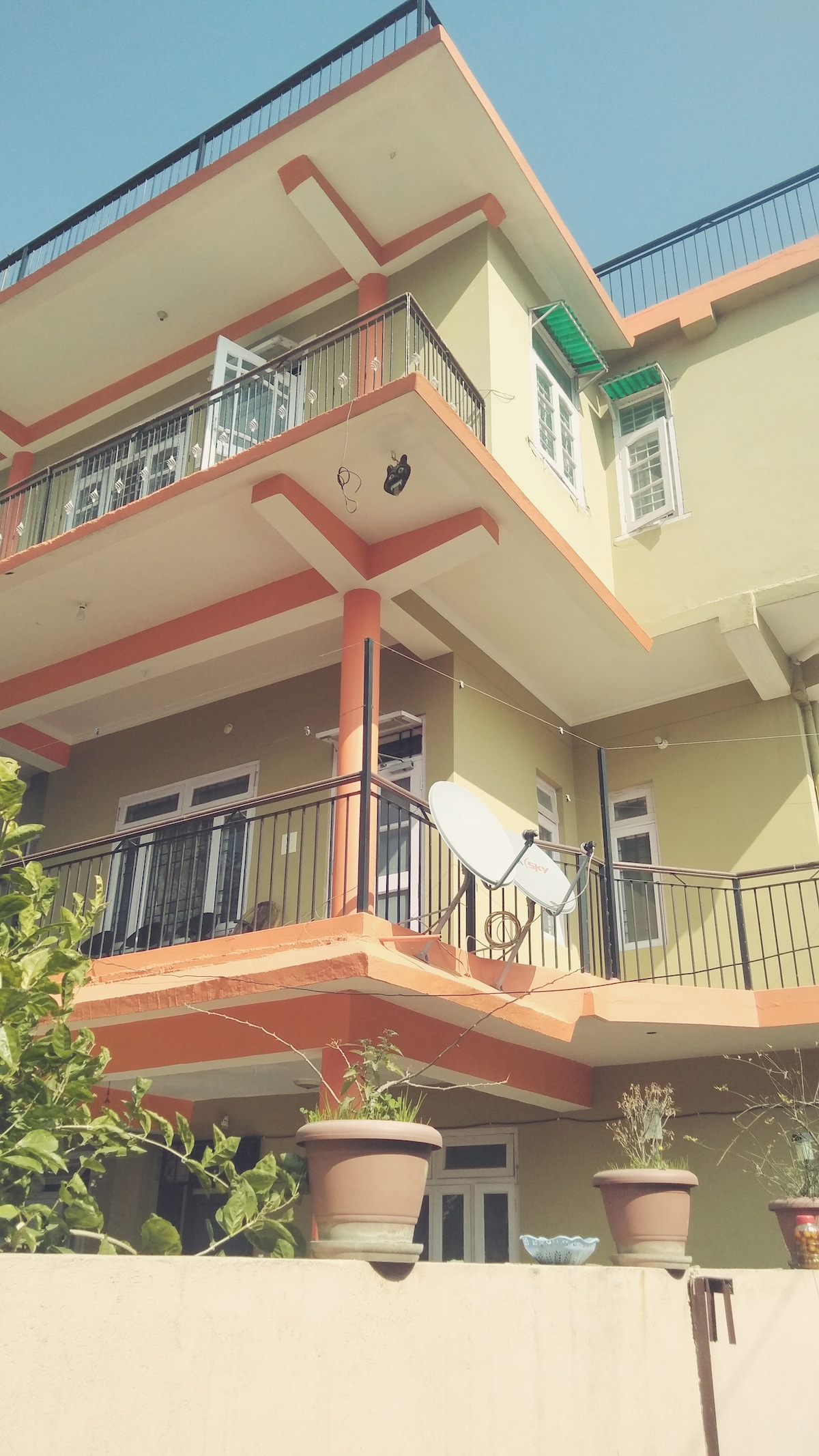 A three-story building features a blend of vibrant yellow and orange exteriors, with balconies adorned by railings. Potted plants are placed along the terrace and ground level, and a satellite dish is mounted on the wall. The clear blue sky provides a bright backdrop.