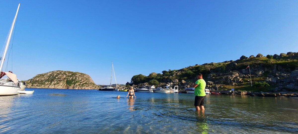 The image captures a serene beach scene with two individuals wading in calm, clear water. Several boats are visible anchored along the shore, while a gentle hillside with greenery provides a natural backdrop under a clear blue sky.