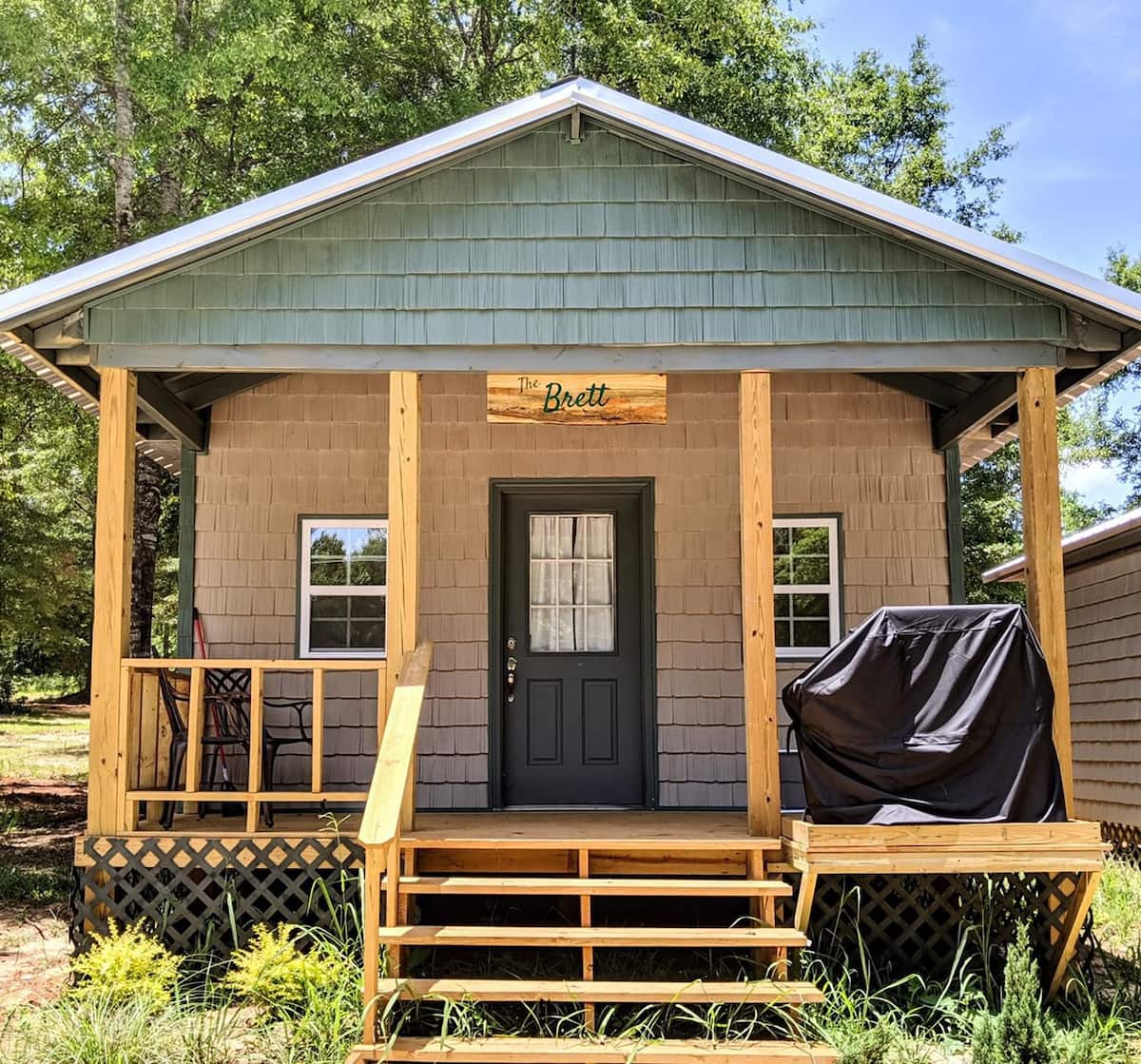 A rustic cabin features a covered front porch with steps leading to the entrance. A wooden sign reads 'The Brett,' and a grill is positioned on the porch. Surrounding greenery provides a natural setting.