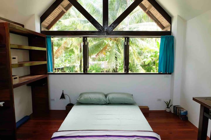 The gallery-style loft bedroom, with large windows, high ceiling, air-conditioning, ceiling fan, queen size bed with luxury spring mattress, open wardrobe, and desk.