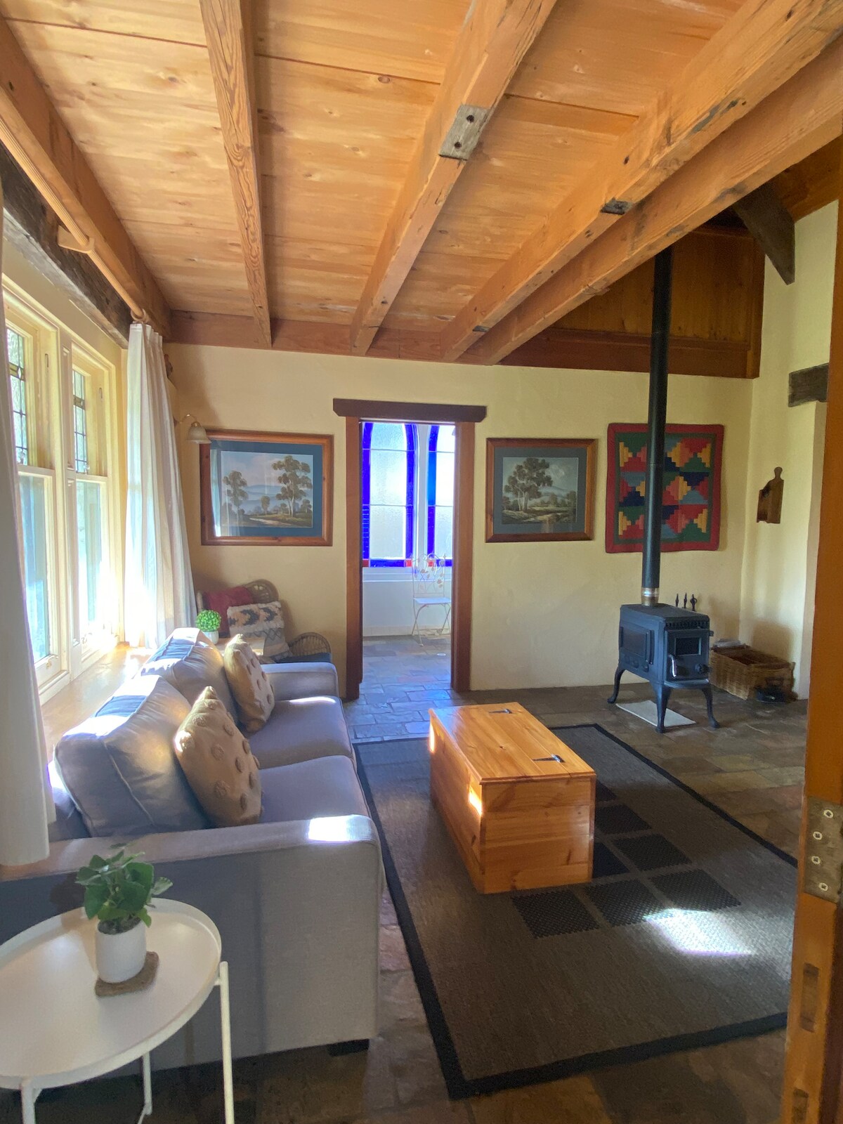 A cozy living area is highlighted by a charcoal sofa adorned with accent pillows. A wooden coffee table sits on a patterned rug, while natural light enters through windows framed in stained glass. A compact wood stove provides warmth, complemented by exposed wooden beams above.