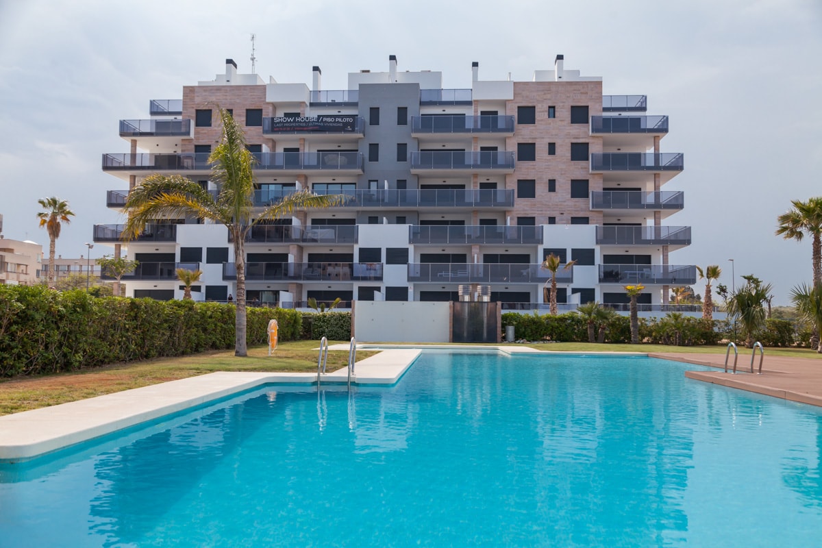 A large swimming pool is set against the backdrop of a modern apartment building. Palm trees are visible around the pool area, providing a touch of greenery. The building's multiple balconies face the pool, enhancing the outdoor space's inviting atmosphere.