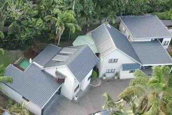 An aerial view of a spacious garden cottage and surrounding structures is presented, showcasing a well-maintained roof and various outdoor areas. The landscape is filled with palm trees and green foliage, providing a serene setting.