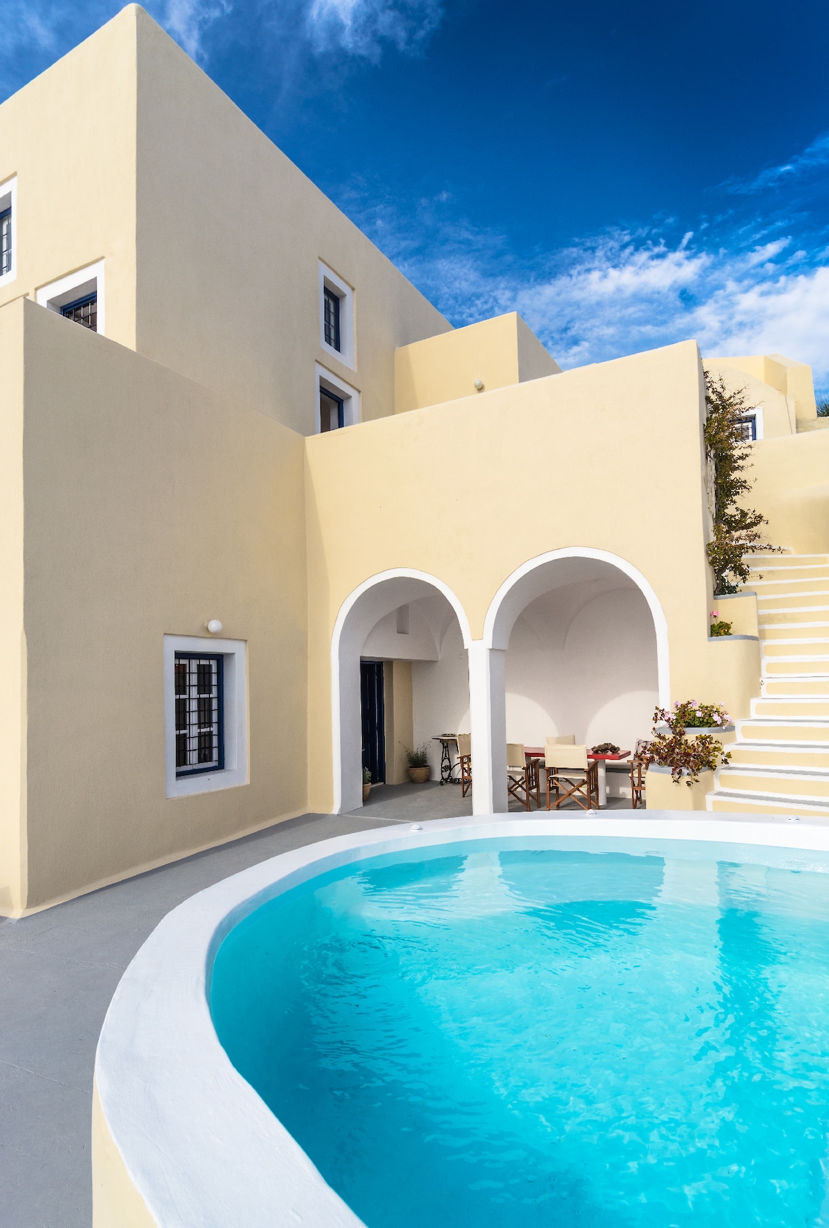 The exterior features a spacious terrace with a circular pool centered in front of a two-story yellow structure. Arched entrances provide access to shaded sitting areas, while stairs lead to upper levels. Clear blue skies add brightness to the inviting atmosphere.