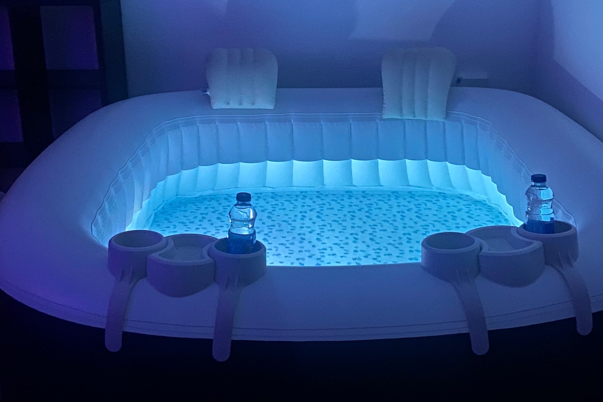 An inflatable jacuzzi is visible, featuring a soft illuminated interior with a soothing blue glow. Two built-in headrests provide comfort, while cup holders are integrated into the design, each holding a bottled water. The overall setup offers a relaxing space for up to six people.