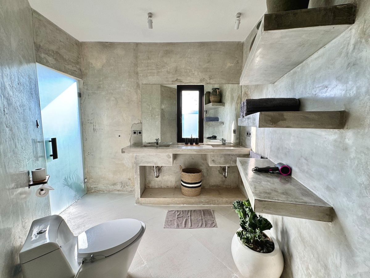 A modern bathroom features polished microcement walls and a minimalist aesthetic. A large vanity with built-in shelves provides storage and decor, while a glass shower is visible to the left. A potted plant adds a touch of greenery, complementing the neutral tones.