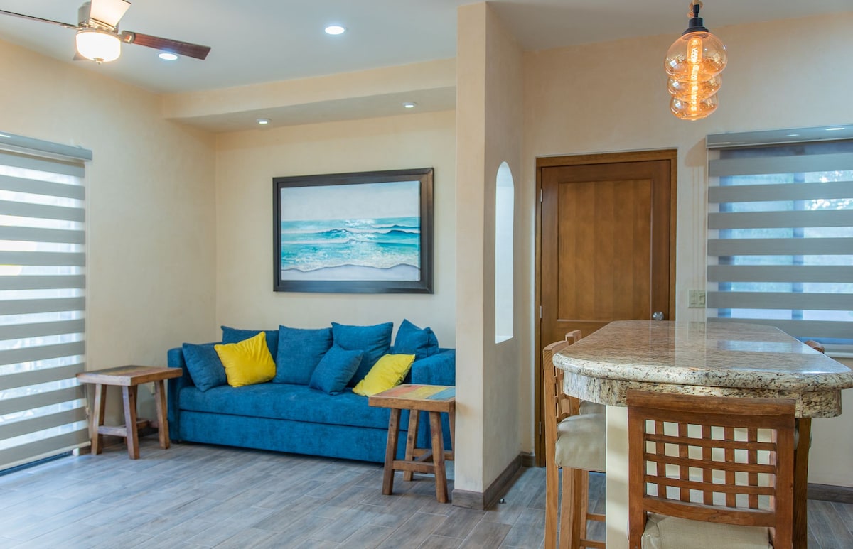 A cozy living area features a blue sofa adorned with yellow cushions, complemented by two wooden side tables. A granite kitchen bar with tall stools is visible, alongside a colorful ocean-themed artwork that enhances the space. Natural light filters through modern window shades.