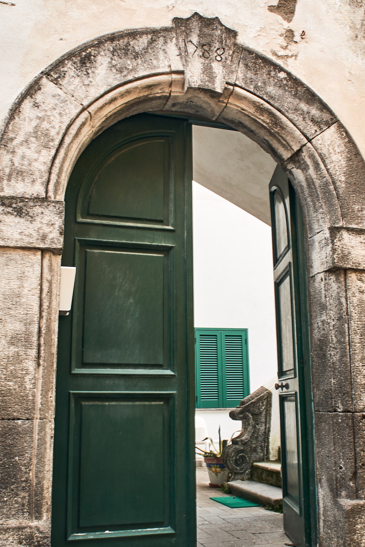 An arched doorway constructed of stone is shown, featuring a dark green wooden door partially open, revealing a glimpse of the interior passage. The year '1888' is engraved at the top of the arch, while green shutters are visible in the background.