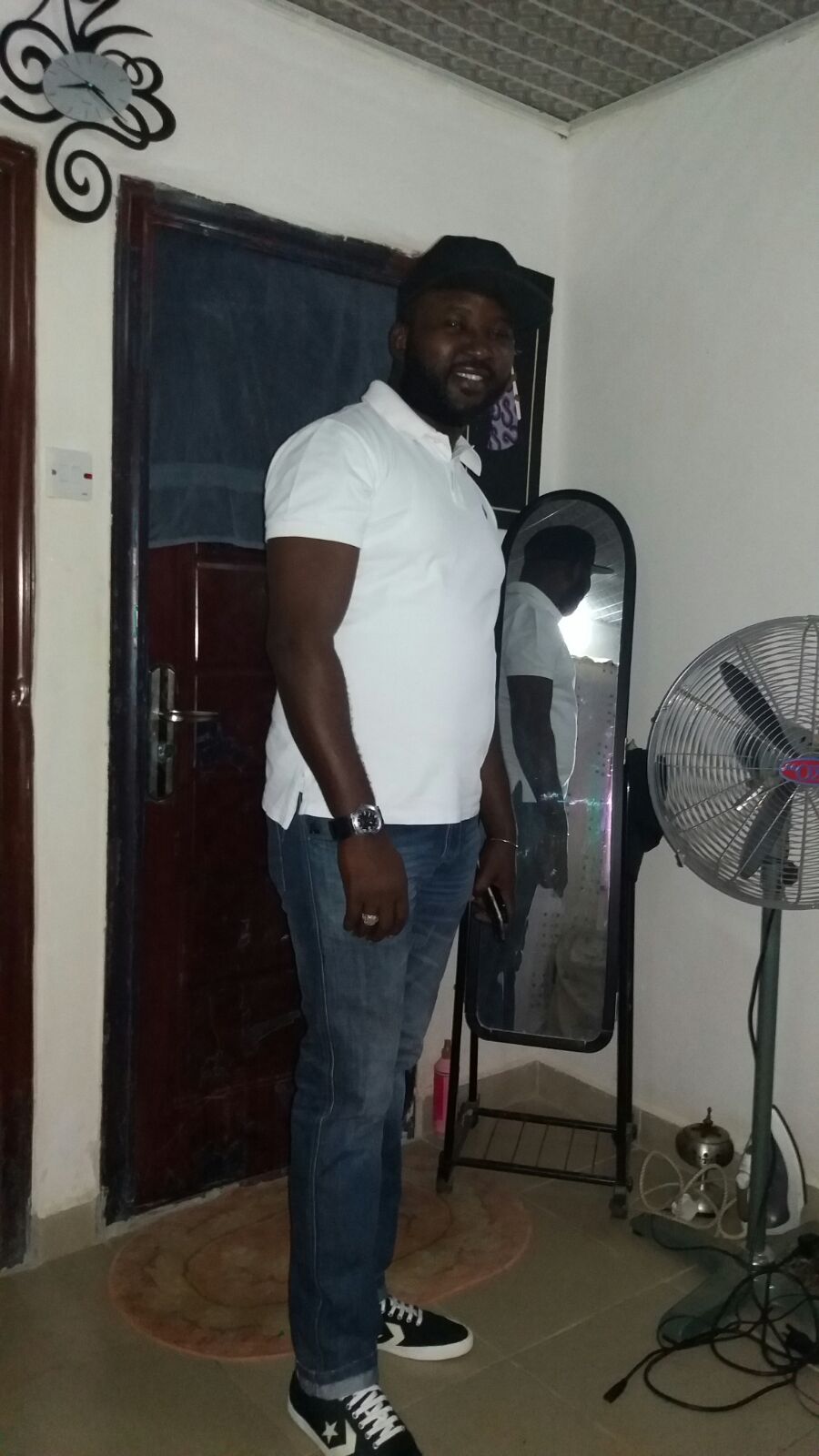 A standing figure in a white shirt and blue jeans is positioned next to a floor mirror on a sleek stand. A fan is visible in the foreground, while a door with a dark curtain and light-colored floor tiles are present in the background.