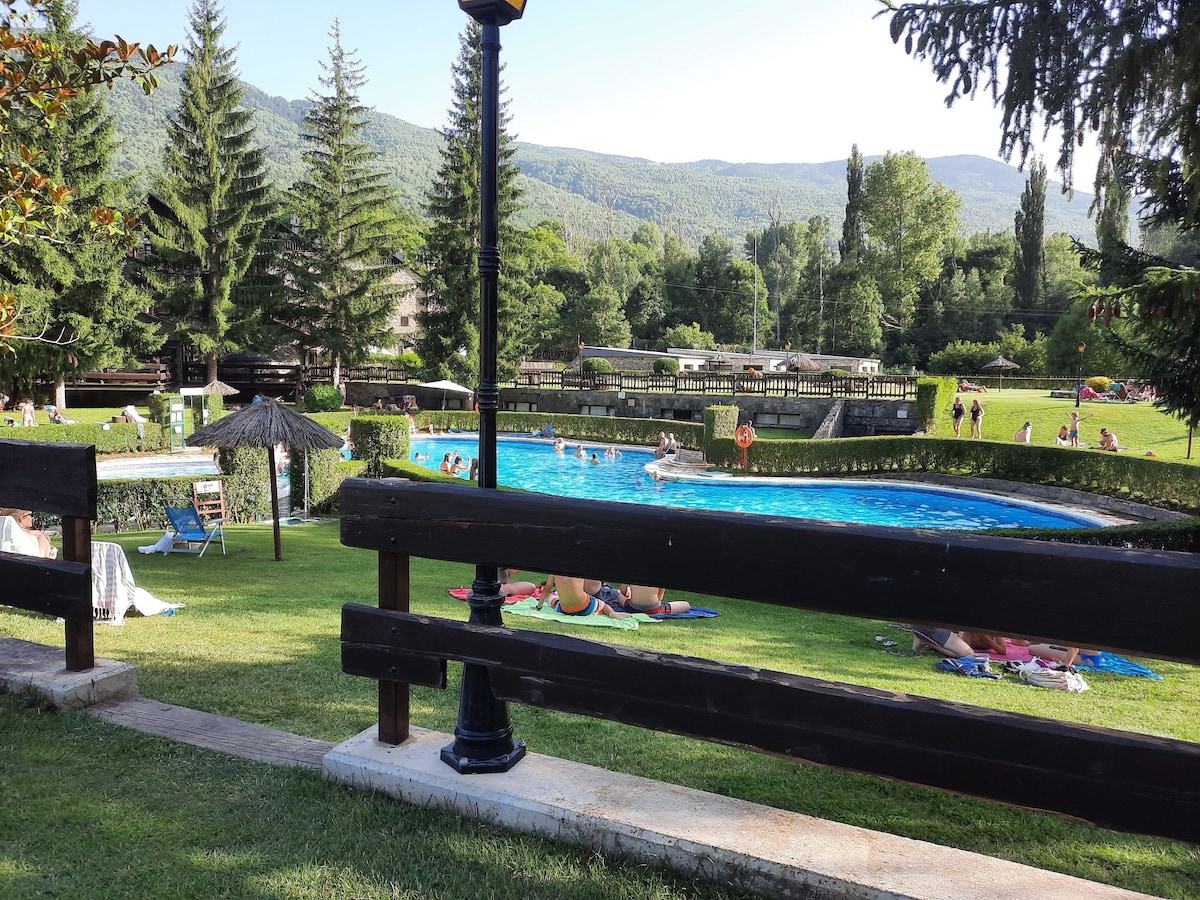 A well-maintained outdoor area is visible, featuring a large swimming pool surrounded by lush green lawns. Umbrella-covered seating areas and sun loungers are arranged on the grass. Scenic hills in the background add to the tranquil environment, while trees provide ample shade.