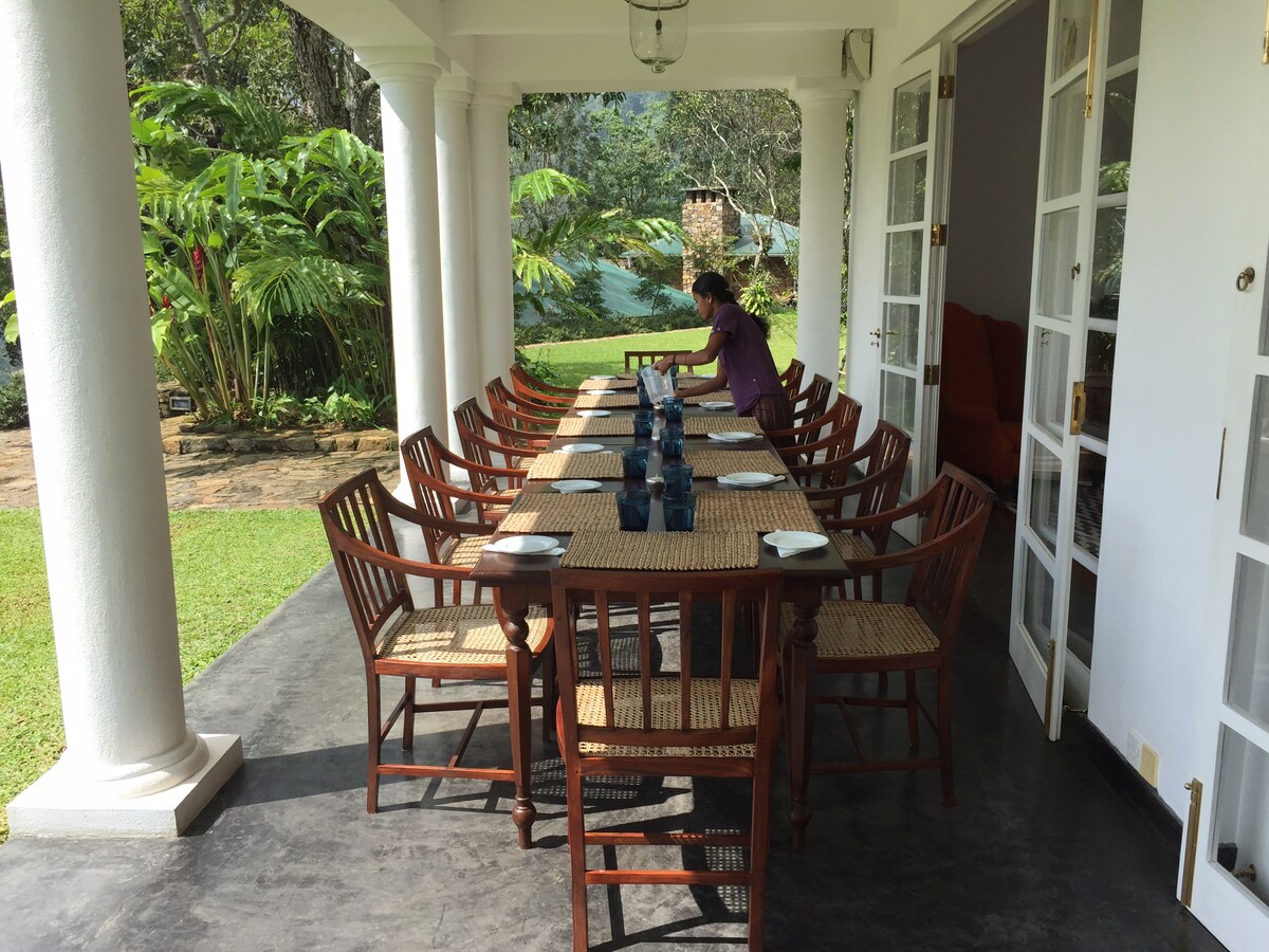 A spacious dining area features a long wooden table surrounded by multiple chairs, set in an open-air veranda. Lush greenery and trees are visible beyond the patio, contributing to a serene environment for meals or gatherings. The floor is made of smooth stone.