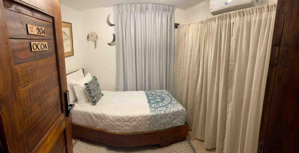 A cozy studio room features a single bed dressed in white linens and a blue patterned throw. Light-colored curtains frame the window, while decorative wall elements add a touch of design. The rustic wooden door stands open, inviting a glimpse of the serene space.