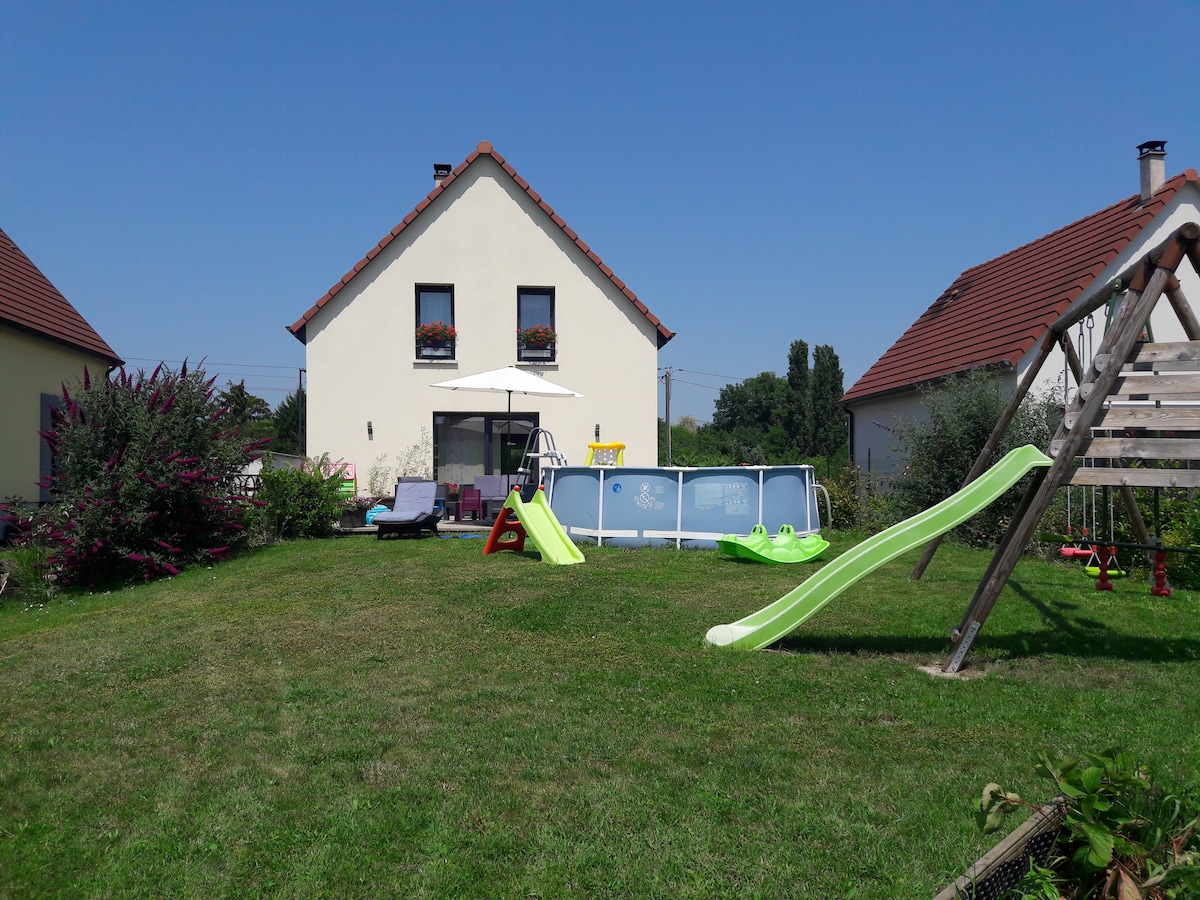 A spacious backyard is showcased, featuring a green lawn with a variety of play equipment. A summer pool is positioned adjacent to the house, which is visible in the background. A swing set, slide, and lounge chairs are organized for outdoor enjoyment.
