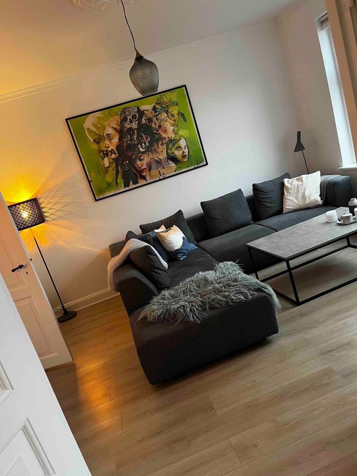 A modern living area is featured with a dark sectional sofa arranged around a square coffee table. A colorful artwork adorns the wall, while a floor lamp and a table lamp provide soft lighting. Light-colored wood flooring adds warmth to the space.