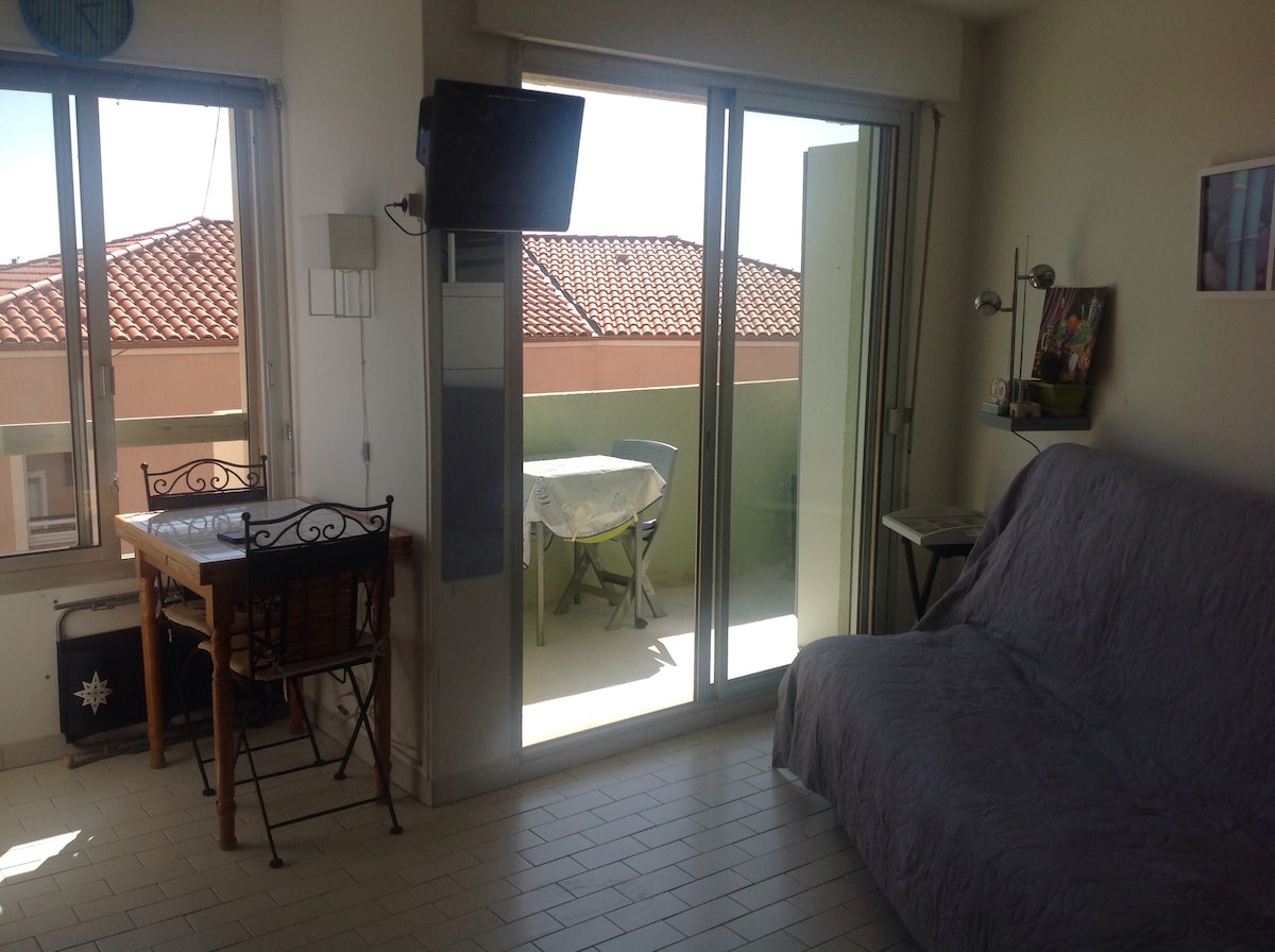A well-lit living area features large glass doors that open to a balcony, allowing natural light to fill the space. A comfortable sofa is positioned beside a small dining table and chairs. A television is mounted on the wall across from the seating area.