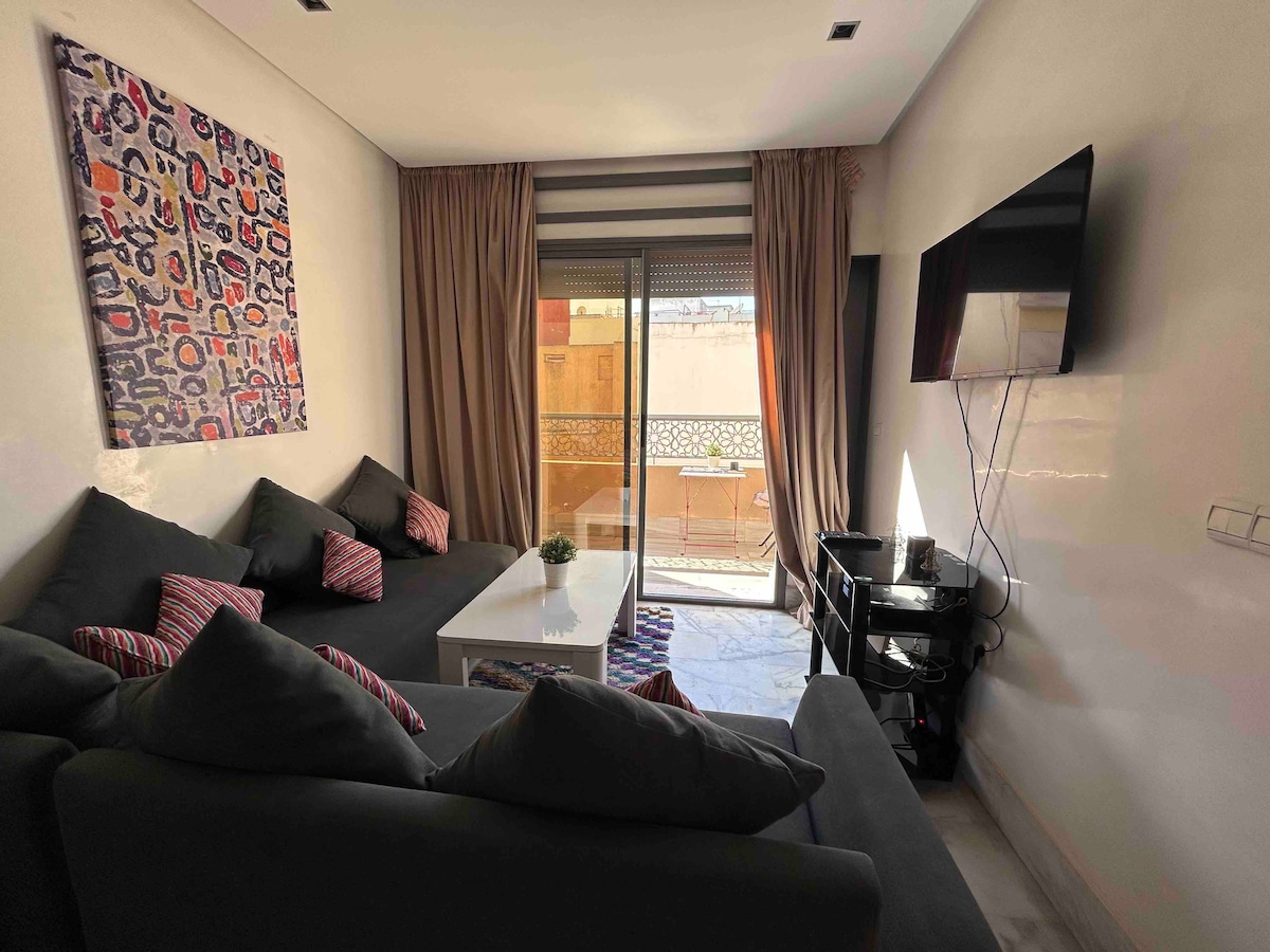 A cozy living area features a dark sofa adorned with colorful cushions, complemented by a sleek white coffee table. Natural light filters through glass doors leading to a balcony, while an abstract art piece adds character to the wall. A television is mounted for entertainment.