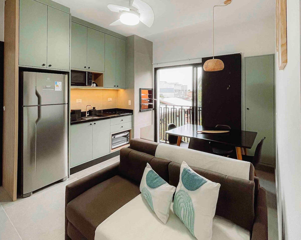 A modern kitchen area features sleek cabinets and stainless steel appliances. A dining table is positioned beside a large window, allowing natural light to brighten the space. A comfortable sectional sofa adorned with decorative pillows offers a cozy spot to relax.