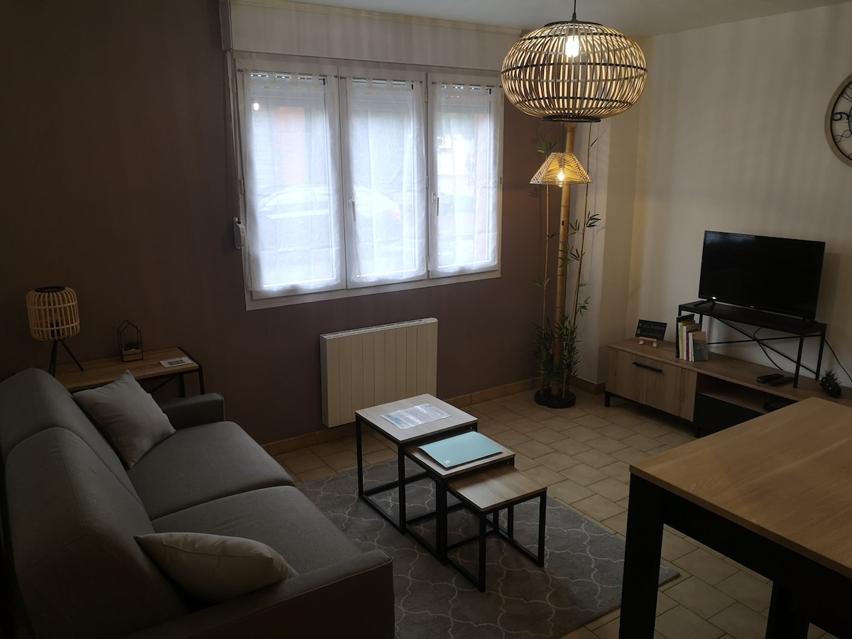 A cozy living area features a spacious sofa positioned across from a television. A set of small tables sits on a patterned rug in front of the seating, with natural light filtering through the large windows dressed in light shades. A floor lamp and decorative plant add warmth.