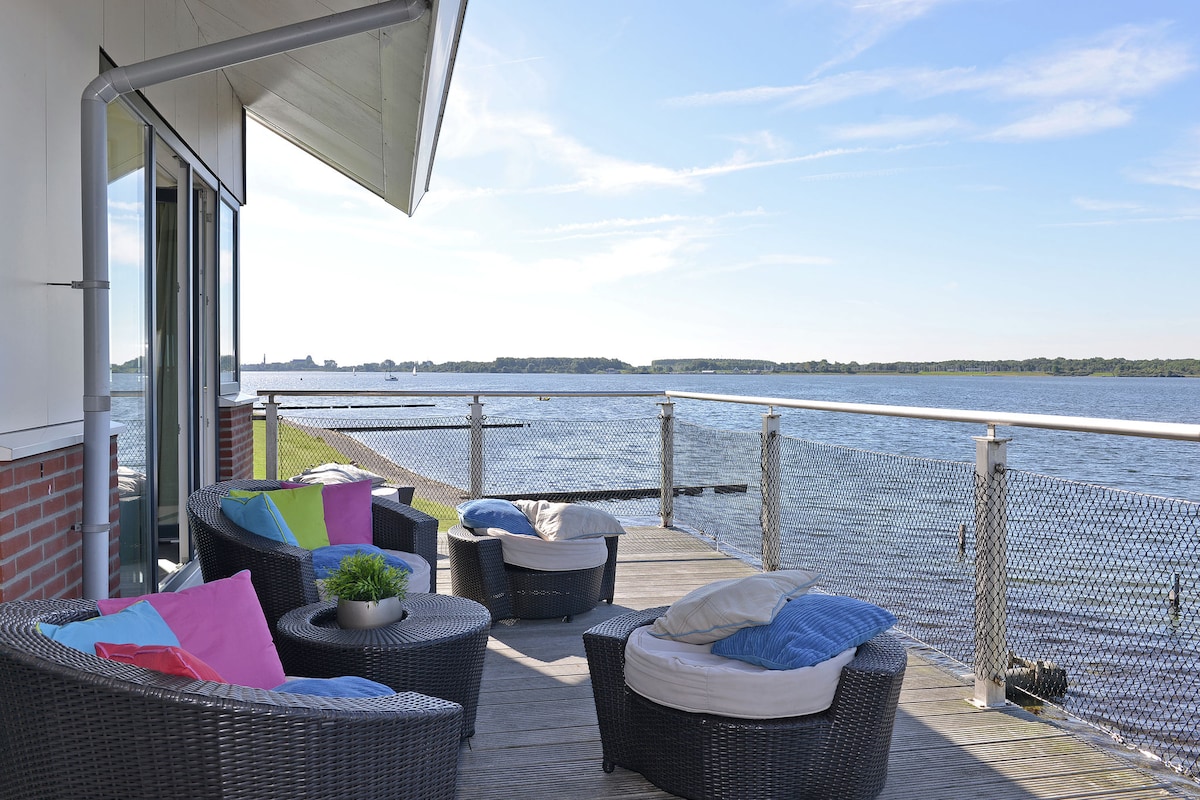A spacious terrace features comfortable seating arrangements with colorful cushions, offering an unobstructed view of the water. The railing, designed with fine mesh, enhances safety while allowing for an expansive vista of the serene lake and surrounding landscape.