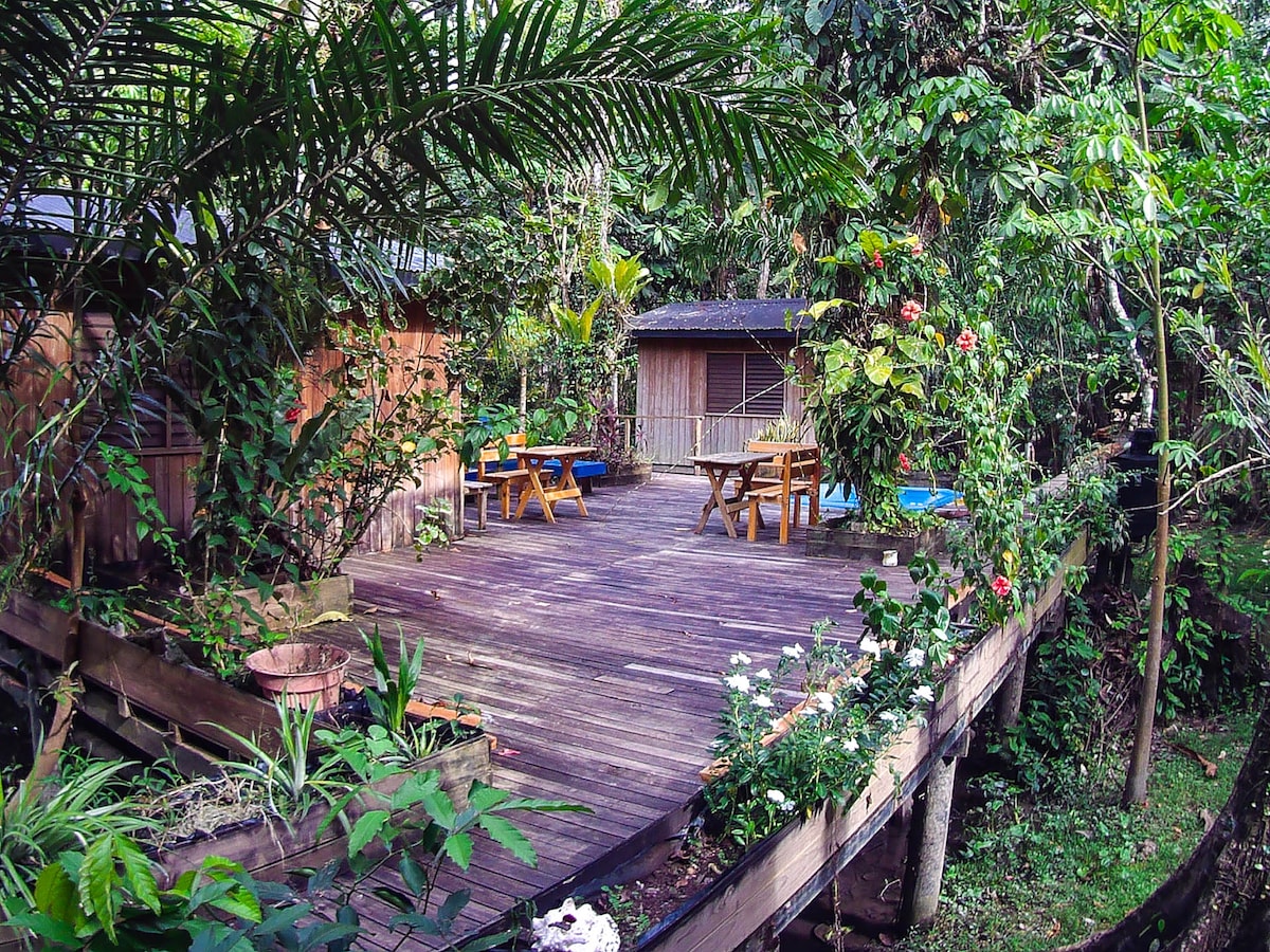 A wooden deck surrounded by vibrant greenery is highlighted, featuring several picnic tables and chairs. Tropical plants and flowers enhance the natural ambiance, creating an inviting outdoor space that encourages relaxation. The rustic buildings in the background are nestled within the lush surroundings.