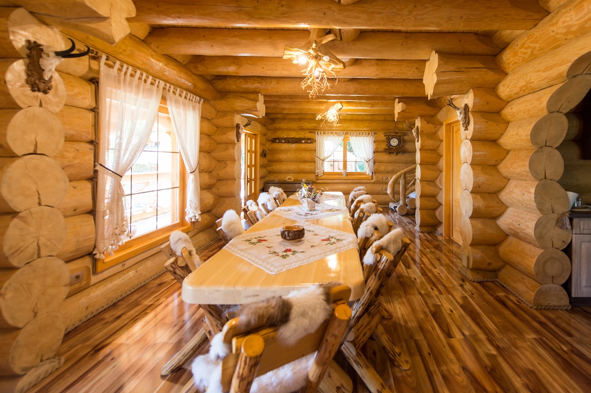 A cozy dining area is highlighted by a long wooden table surrounded by rustic chairs, each adorned with soft textures. Large windows allow natural light to fill the space, and log walls add warmth to the inviting atmosphere.