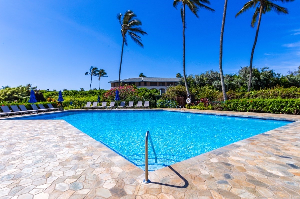 A modern pool is surrounded by a stone deck and lounge chairs. Lush greenery and palm trees frame the area, creating a serene atmosphere. The clear water reflects the blue sky overhead, inviting relaxation and enjoyment.