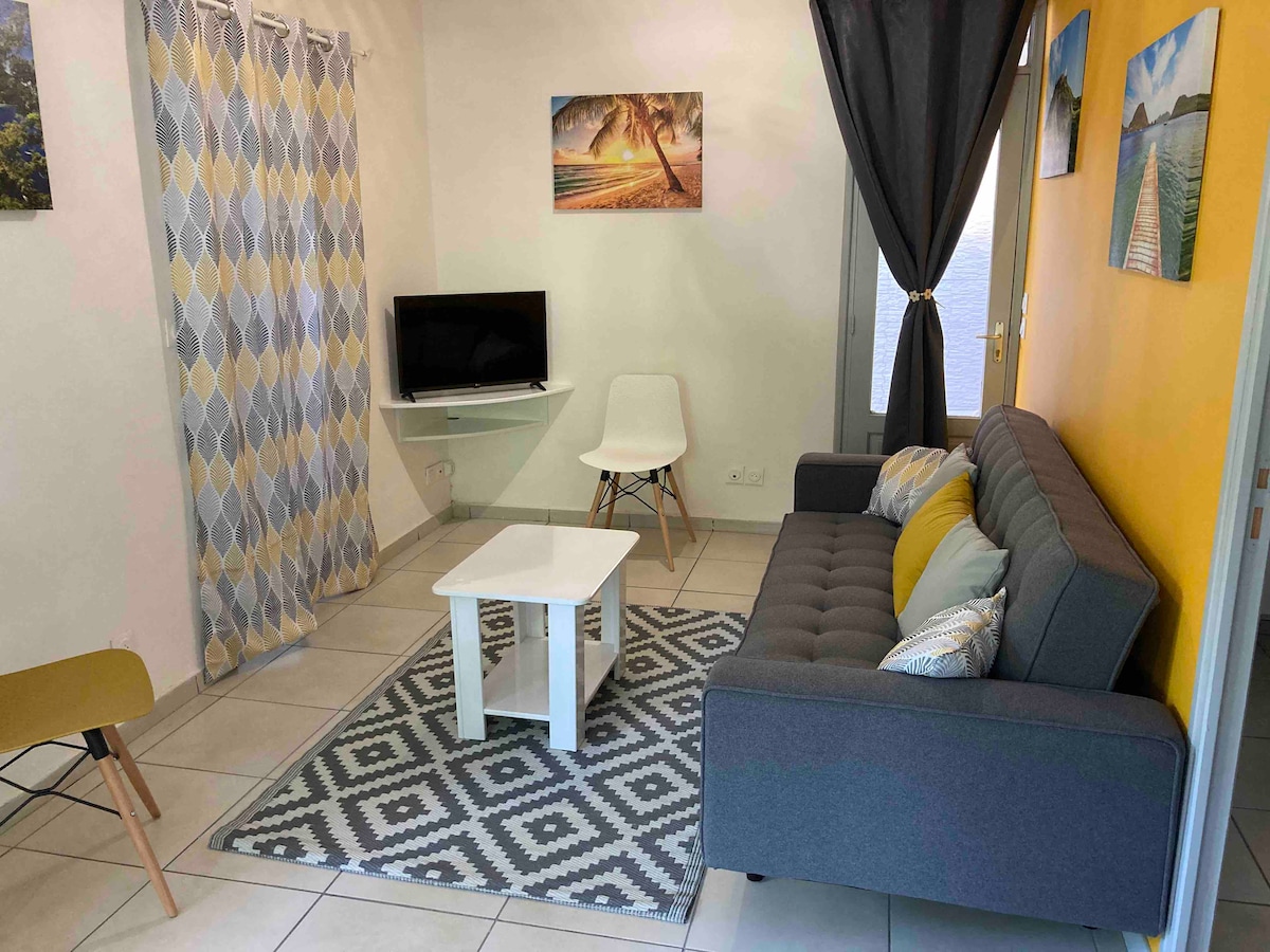 A cozy living area features a gray sofa positioned across from a small white coffee table. A television is mounted on the wall, surrounded by artwork depicting tropical scenes. Light enters through a window dressed with dark curtains, enhancing the bright yellow accent wall.