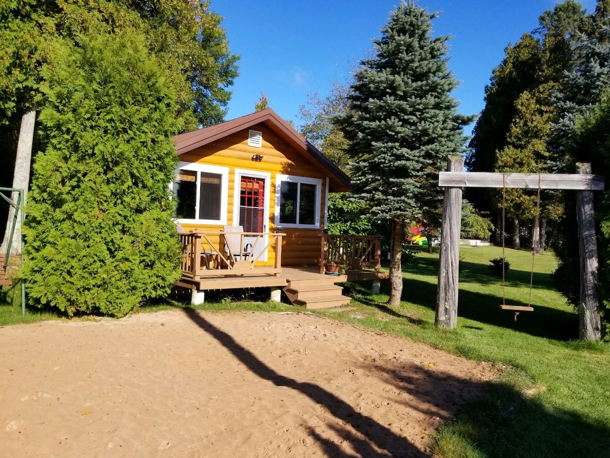 A charming cottage with a yellow exterior is framed by tall trees and shrubs. A sandy area is visible in the foreground, leading up to a raised porch with steps. A swing dangles from a wooden frame beside the cottage, enhancing the outdoor space.