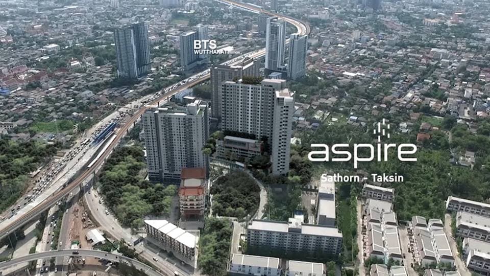 An aerial view showcases the Aspire condo complex situated on Sathorn-Taksin in proximity to Wutthakat BTS. Surrounding urban landscape features a mix of high-rise buildings and residential areas, with roadways visible connecting different parts of the city.