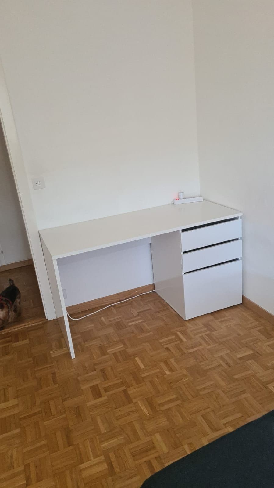 A simple white desk is positioned against a wall, complemented by three drawers for storage. The floor features a warm wooden parquet pattern, contributing to the space's calm aesthetic.