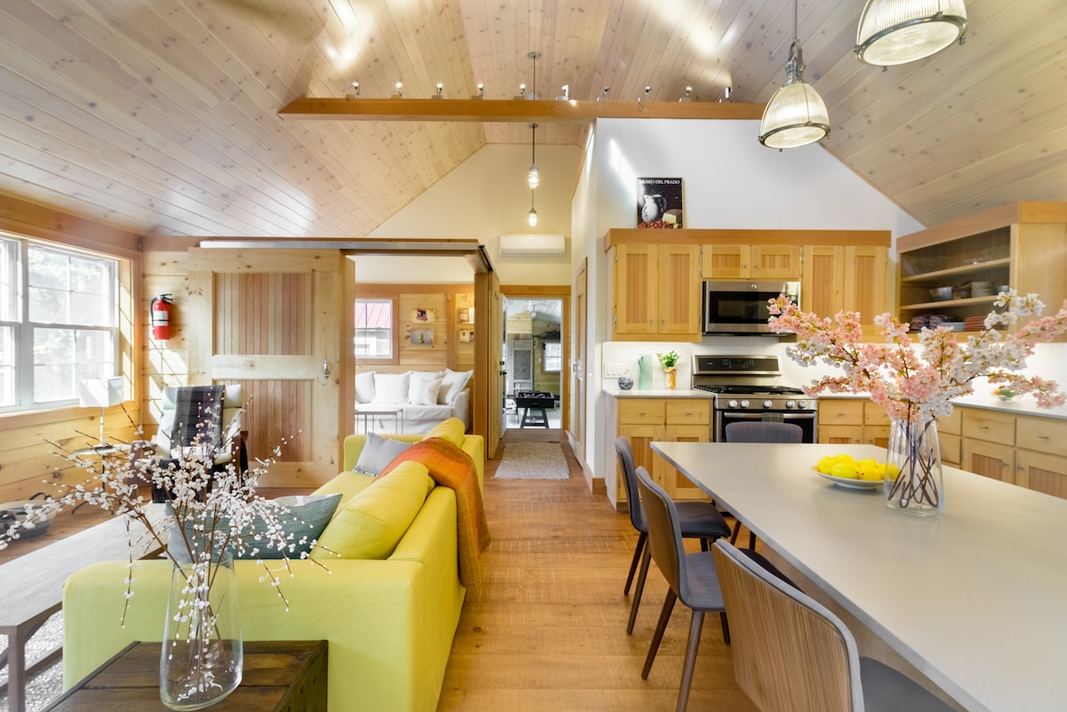 An open-concept living area features warm wood paneling and high ceilings, creating a bright and inviting space. A modern kitchen is visible, equipped with stainless steel appliances, while a dining table is set with fresh fruit. A colorful couch and decorative floral arrangements add a touch of comfort.
