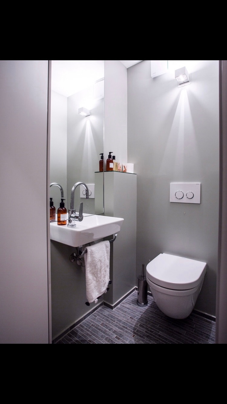 A modern bathroom is illuminated by soft wall sconces. A compact sink with a sleek faucet is mounted on the wall, accompanied by neatly arranged toiletries. A white toilet features a dual flush system, and the floor is covered with textured tiles.