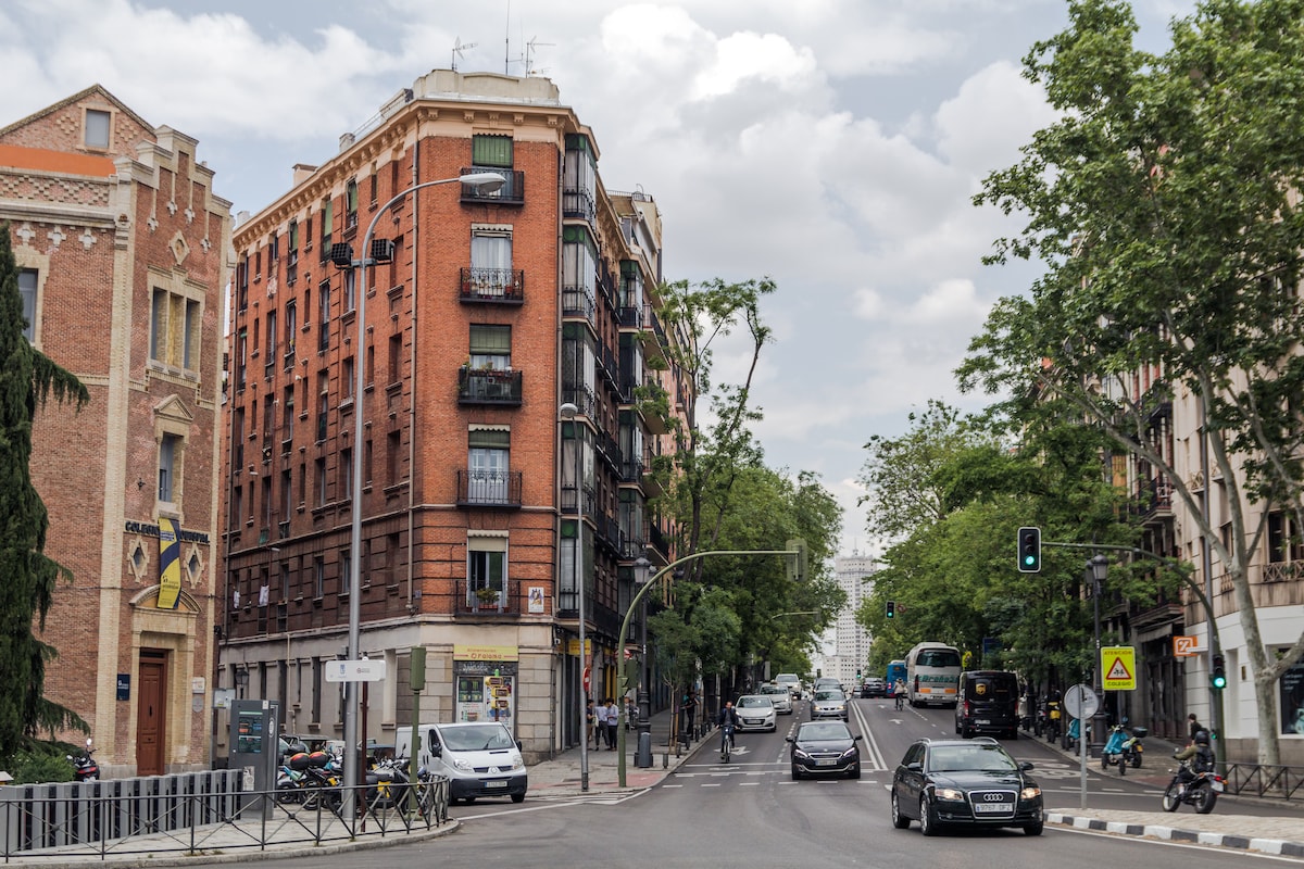 An urban street scene is depicted, showing a brick building with balconies at the corner of a tree-lined avenue. Vehicles are seen on the road, with traffic lights and signs visible. The sky is partly cloudy, enhancing the city atmosphere.