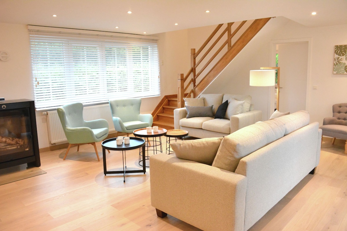 A spacious living area is furnished with a light-colored sofa and two green accent chairs. A small black table sits in front, while a decorative lamp provides soft lighting. Large windows allow natural light to fill the room, highlighting the wooden flooring and staircase.