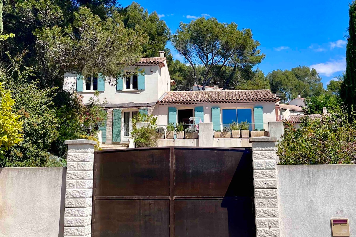 A spacious villa is featured behind an imposing brown gate, with light-colored stone walls surrounding the property. The architecture showcases charming blue shutters and a tile roof. Lush greenery from the surrounding trees adds to the tranquil setting, under a clear blue sky.