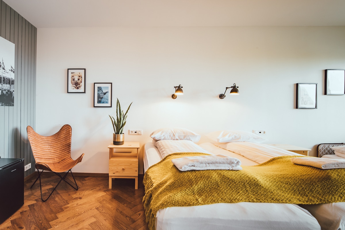 A bright private room features a spacious bed that can be arranged as a double or two singles. A cozy chair and larger plants are positioned beside the bed. Two wall-mounted lamps provide illumination, while framed artworks add a subtle decorative touch.