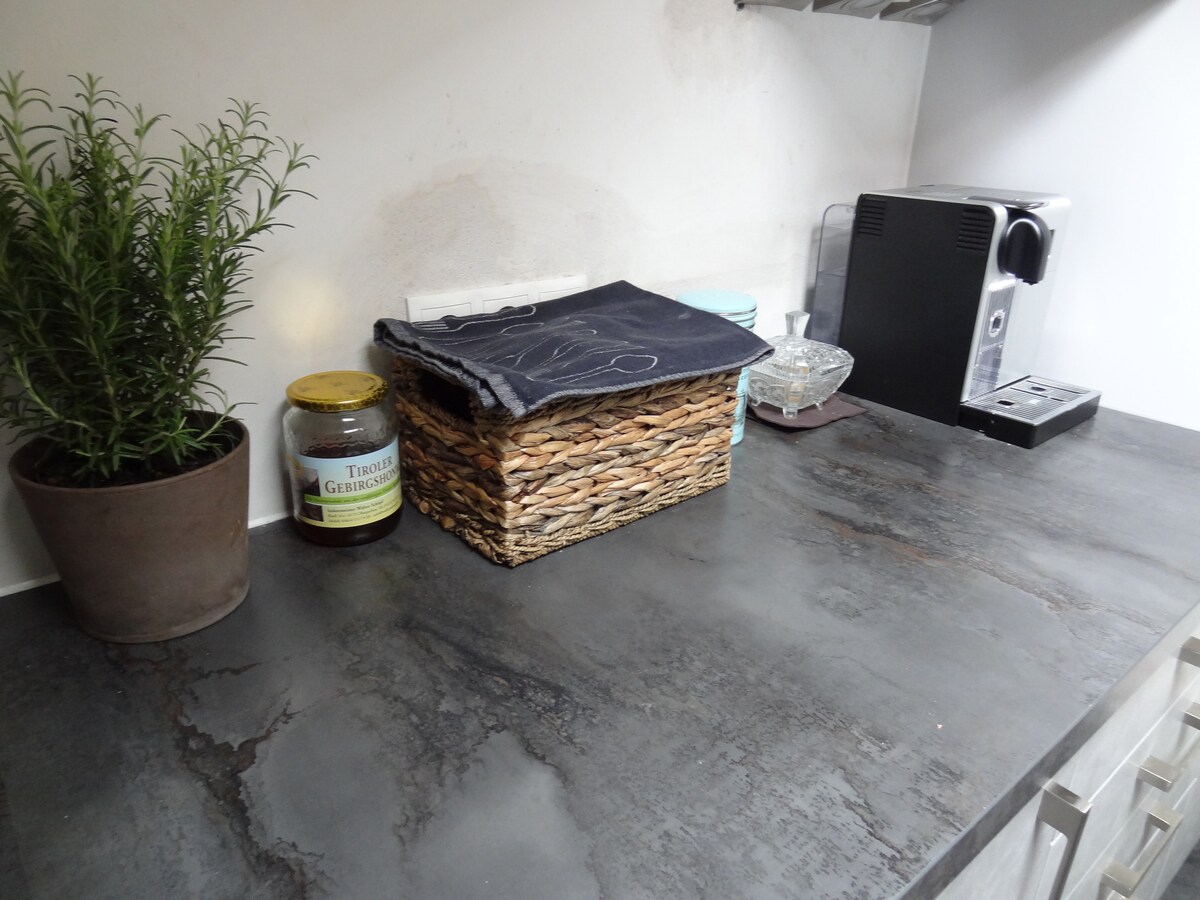 A modern kitchen countertop is displayed, featuring a potted herb, a woven storage basket, a jar of local honey, and an espresso machine. The surface appears sleek and dark, with subtle variations in texture and color.