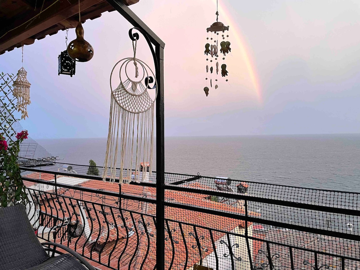 A private balcony displays intricate wind chimes and a decorative macramé hanging, overlooking the sea. A soft rainbow is visible in the distance, framed by a tranquil sky. Comfortable seating is arranged for enjoying the expansive water views.