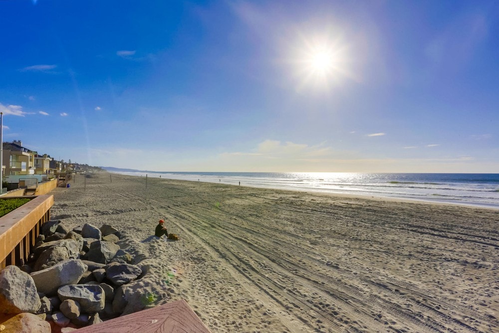 The image captures a wide view of a sandy beach under a clear blue sky. Sunlight reflects off the ocean water, creating a shimmering effect. In the foreground, large rocks border the beach while a solitary figure sits on the sand, enjoying the serene atmosphere.