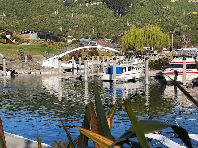 A serene marina view is presented, featuring a white bridge arching gracefully over calm waters. Several boats are docked at wooden berths, with greenery framing the scene. Reflections of the surroundings ripple gently on the water's surface, enhancing the tranquil atmosphere.