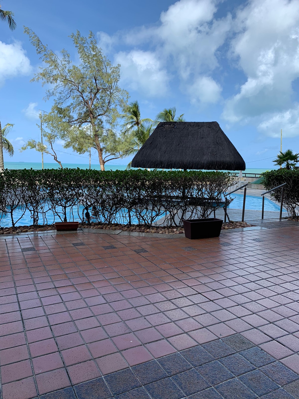 An outdoor pool area is visible, featuring a sun shelter with a thatched roof. Lush greenery surrounds the pool, providing a natural barrier. The pool reflects the clear blue sky, and a smooth, tiled surface leads to the water's edge.