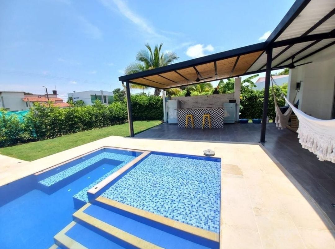 An outdoor area features a swimming pool with steps leading into shallow water, surrounded by a tiled deck. A shaded patio area contains a barbecue station with patterned tiles. Lush greenery frames the space, and two hammocks are visible, ready for relaxation.